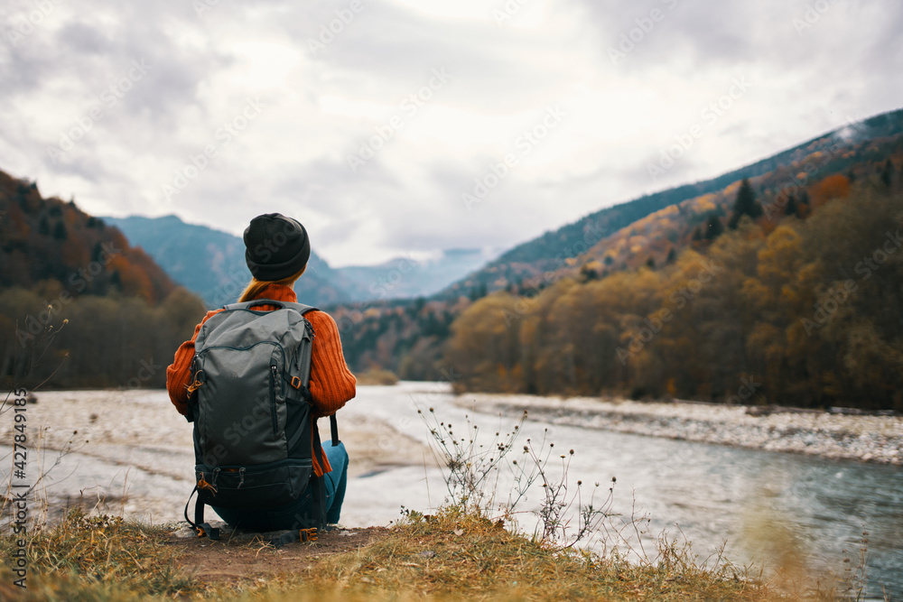 woman with a backpack and in a jacket with a hat on the river bank in the mountains in nature