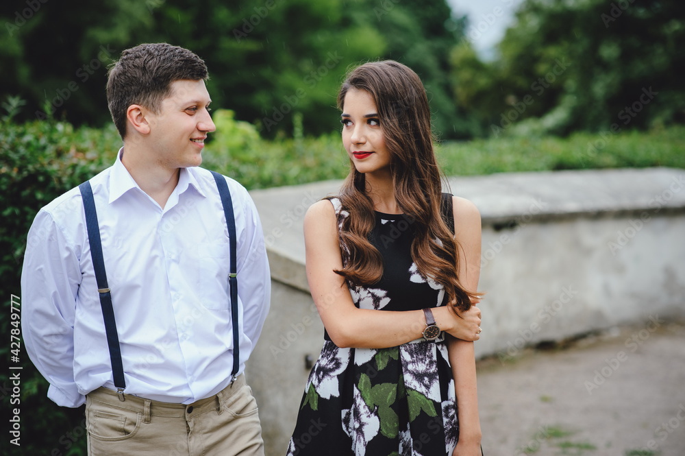 Loving happy couple. Beautiful brunette in dress and young man in white shirt, pants with suspenders walking through city streets. Portrait of a couple in love 