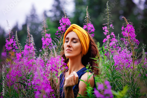 A young, calm woman with dark hair and a turban, eyes closed with happiness, clings to her tall stems of spray with pink flowers in a flower field. Kirei. Willow-grass meadow. Fireweed. photo