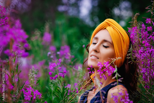 A young, calm woman with dark hair and a turban, eyes closed with happiness, clings to her tall stems of spray with pink flowers in a flower field. Kirei. Willow-grass meadow. Fireweed. photo