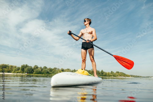 Athletic muscular man with a deaf torso stands on a sup board with an oar in his hand and looks away with a serious face.