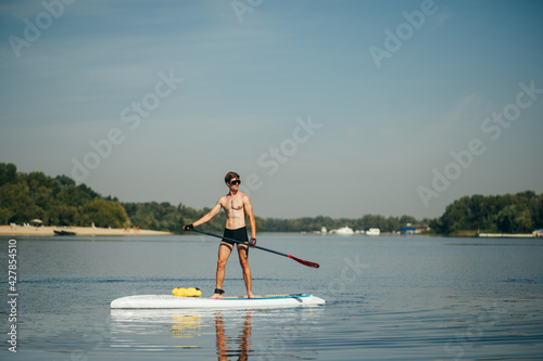 Athletic young man paddles standing on a sup board on the river and looks to the side. A muscular man is active on the water.