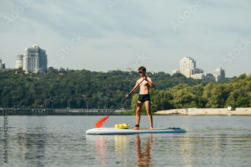 Muscular young man stands on sup board on water and paddles on background of beautiful landscape. Fitness on sup board © bodnarphoto