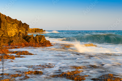 Turquoise sea with waves and splashes and a rocky orange beach on a summer evening