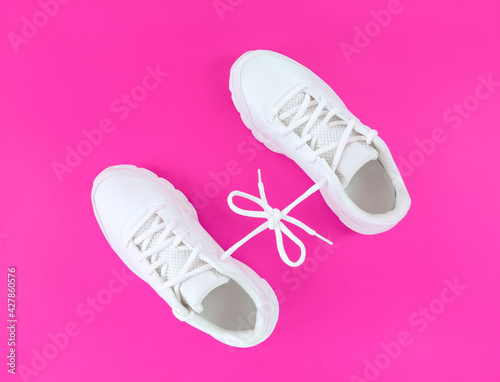 Pair of white sport shoes connected with laces bow on pink background.