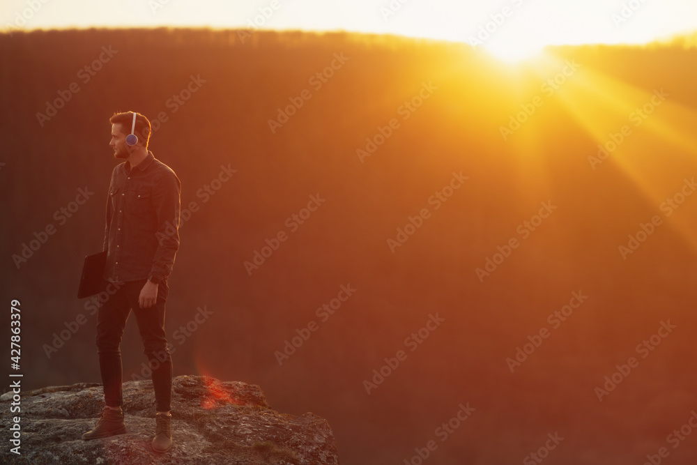 Man with headphones standing on the rocky mountain with pine forest and sunset on the background.