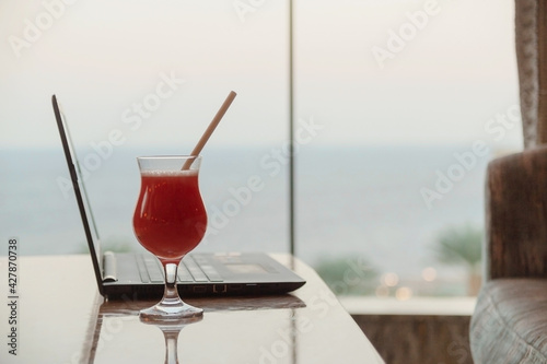 A strawberry cocktail next to a laptop in a bar overlooking the sea. Remote work on vacation concept. © svitlini