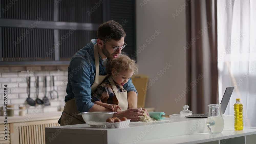 dad and toddler son are cooking by internet recipe, looking on laptop and making dough on kitchen table, father and baby boy