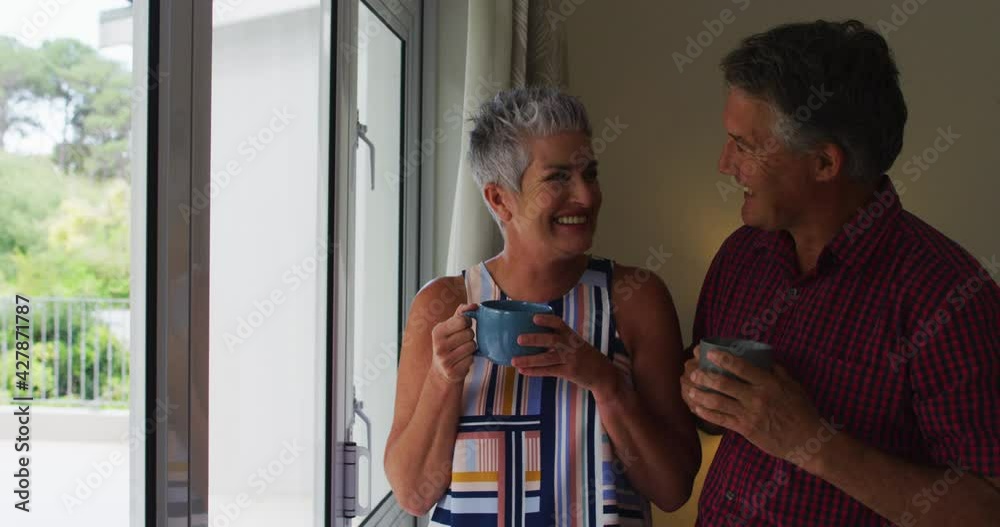Caucasian senior couple smiling and drinking coffee together while looking out of the window at home Caucasian senior couple smiling and drinking coffee together while looking out of the window at home