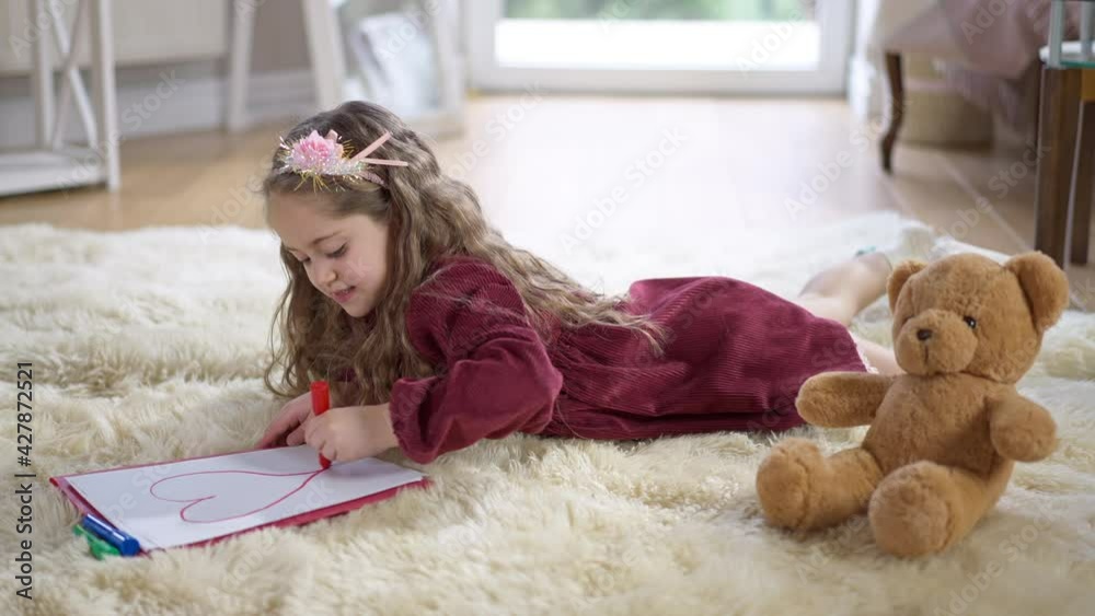 Concentrated curly-haired cute girl drawing red heart on paper lying on soft carpet in living room. Wide shot side view of relaxed Caucasian pretty child enjoying leisure at home indoors Concentrated curly-haired cute girl drawing red heart on paper lying on soft carpet in living room. Wide shot side view of relaxed Caucasian pretty child enjoying leisure at home indoors
