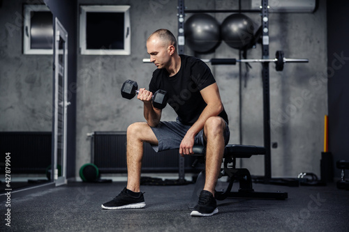 Pumping arm and shoulder muscles. A man in a black T-shirt lifts a dumbbell with one hand while sitting in the gym. Strengthening the triceps and biceps. Sports lifestyle and fitness challenge