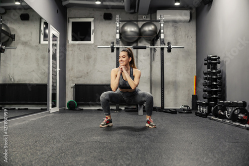 Squatting and burning the muscles of the buttocks and legs. Portrait of a hot woman in sportswear and good physical shape doing squats in an isolated indoor gym. Strength and motivation, fitness goal
