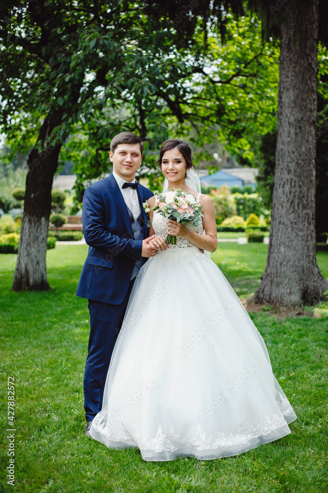 The groom in a blue tuxedo is hugging the bride in a white dress with a mesh. Beautiful wedding bouquet of pink and white peonies in the hands of the bride.