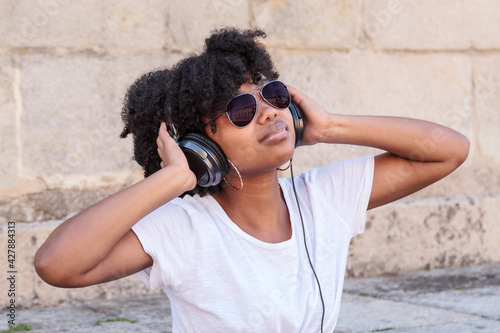 Young black woman with afro hair listens to music with her headphones and sunglasses outdoors on a hot summer day