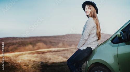 Young beautiful stylish woman in hat standing next to her car traveling in the countryside overlooking the hills