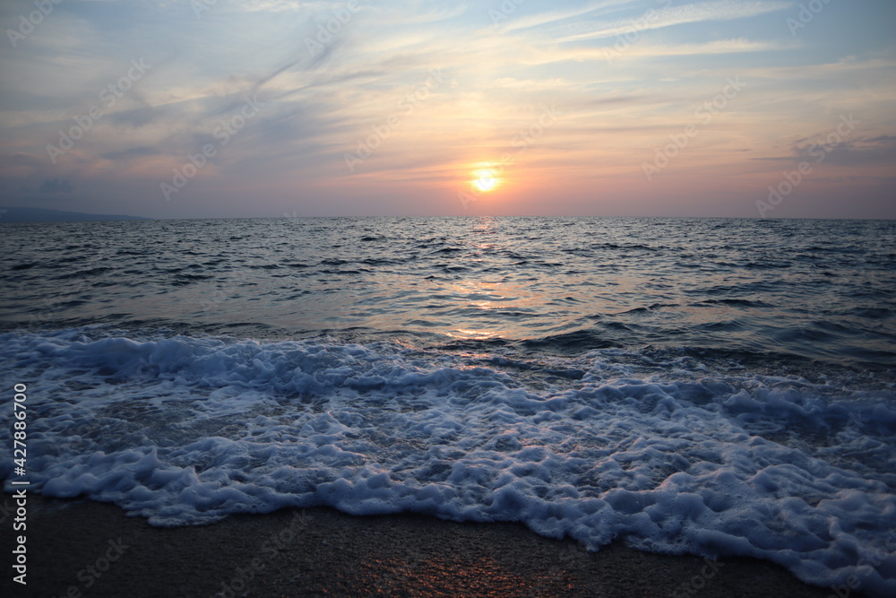 Fototapeta premium Scenic sunset by the sea in pastel colors, Calabria, Italy. Seascape sun on the horizon reflected in waves and sea foam at sunset on a summer evening