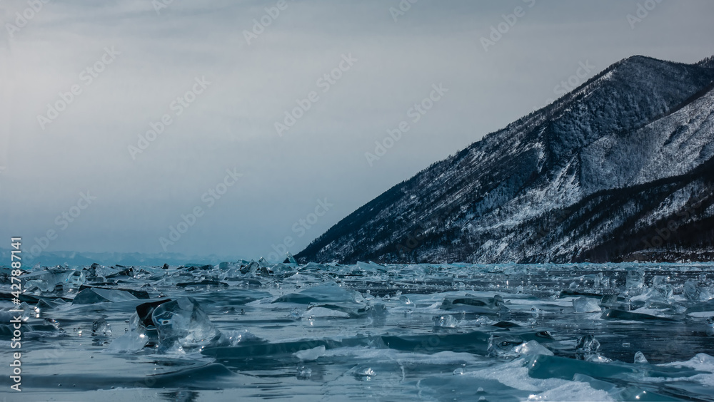 Obraz premium Transparent shiny ice floes are scattered over the surface of the frozen lake to the horizon. Against the background of the sky, there is a wooded and snow-covered mountain. Pastel shades. Baikal