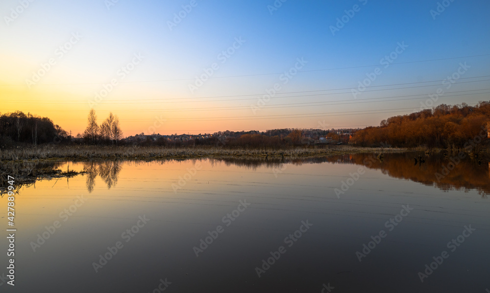 Golden sunset on the lake with trees and reeds in the background. Townhouses and huts lit by sun in the distance.