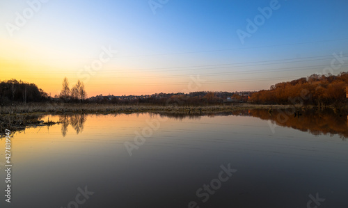 Golden sunset on the lake with trees and reeds in the background. Townhouses and huts lit by sun in the distance.