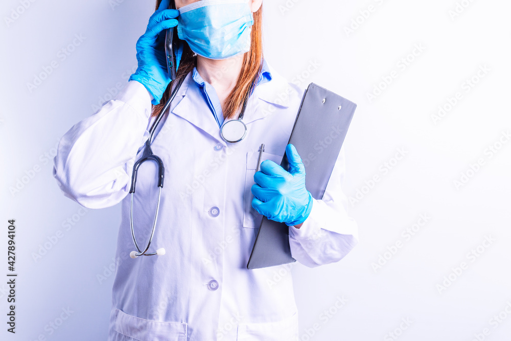 Caucasian young woman wearing stethoscope gown and gloves and mask holding mobile phone to one ear and reports in the other. technology and medicine communication concept