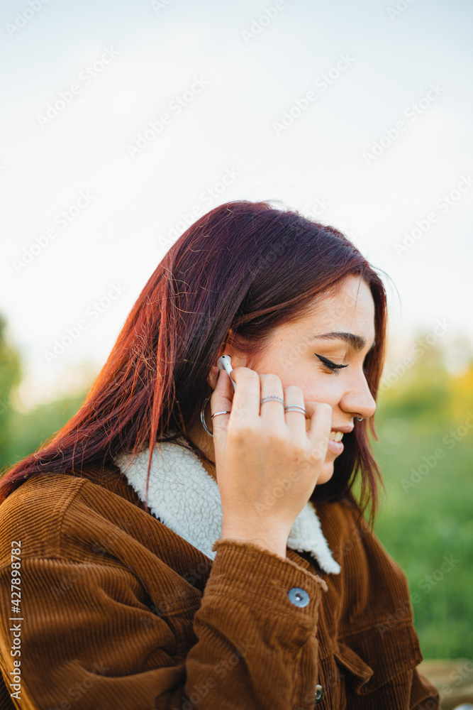 A teenager putting a white wireless ear-phone into her ear while sitting on the grass during sunset A teenager putting a white wireless ear-phone into her ear while sitting on the grass during sunset