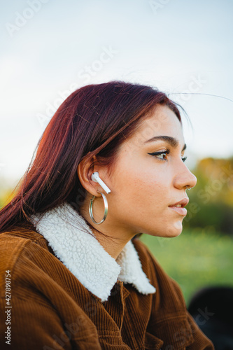 A teenager girl listening to music through white wireless ear-buds. She is sitting on the grass during the sunset.