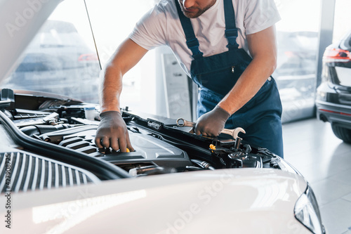Quality service. Young man in white shirt and blue uniform repairs automobile
