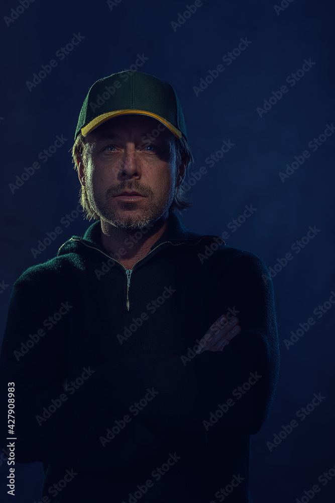 Shaded dark portrait of a blond man with stubble beard and baseball cap in a dark green wool sweater in front of a dark blue wall. Shaded dark portrait of a blond man with stubble beard and baseball cap in a dark green wool sweater in front of a dark blue wall.