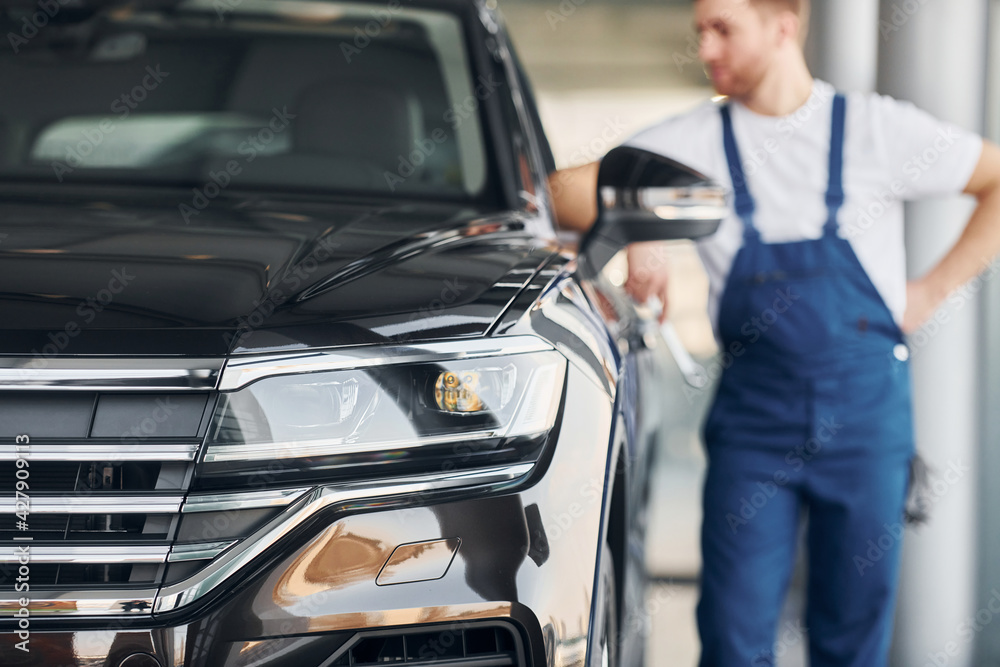 Front view of the car. Young man in white shirt and blue uniform repairs automobile Front view of the car. Young man in white shirt and blue uniform repairs automobile