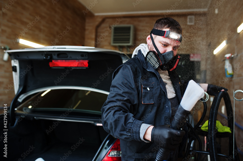 Man in uniform and respirator, worker of car wash center, cleaning car interior with hot steam cleaner. Car detailing concept. Man in uniform and respirator, worker of car wash center, cleaning car interior with hot steam cleaner. Car detailing concept.