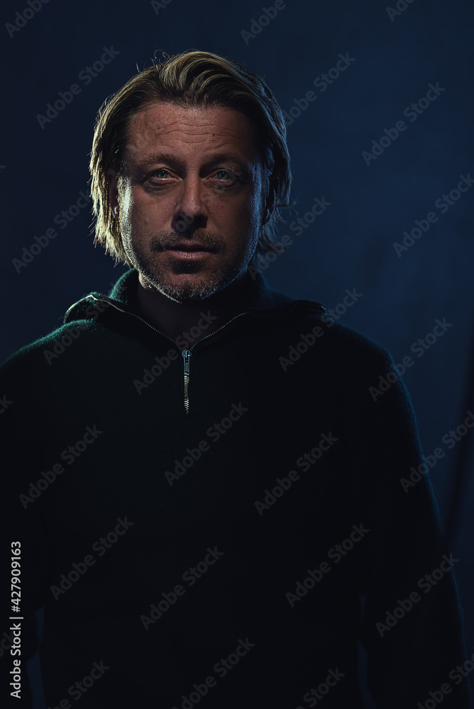 Shaded dark portrait of a blond man with stubble beard in a dark green wool sweater in front of a dark blue wall. Shaded dark portrait of a blond man with stubble beard in a dark green wool sweater in front of a dark blue wall.