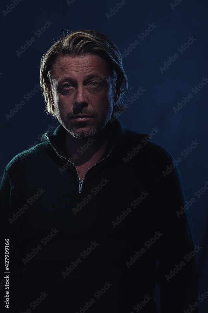 Shaded dark portrait of a blond man with stubble beard in a dark green wool sweater in front of a dark blue wall. Shaded dark portrait of a blond man with stubble beard in a dark green wool sweater in front of a dark blue wall.