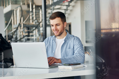 Uses laptop. Young man in white shirt and blue jacket works indoors