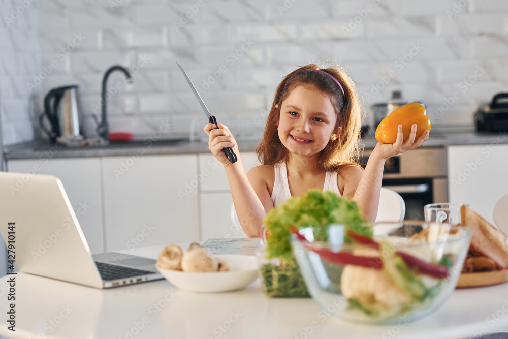 Happy little girl sits on the kitchen with food and laptop on the table