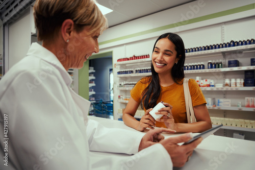 Happy senior female pharmacist showing screen of digital tablet to young customer holding bottle of supplements in pharmacy while in a conversation