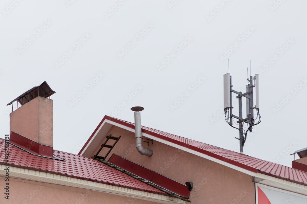 Telecommunication antenna on the roof, cellular and telephone communications. Smoke and ventilation pipes against the gray sky. Red tiles on the roof of a residential building.