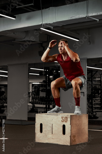 Sexy muscular men using the platform for his legs on a dark colorful background of gym. © Georgii