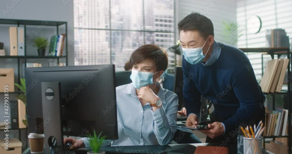 Two colleagues, Asian man and Caucasian woman, communicate in office in protective medical masks, comparing data on their devices. Two colleagues, Asian man and Caucasian woman, communicate in office in protective medical masks, comparing data on their devices.