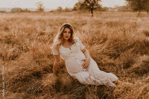 Caucasian pregnant young blonde woman in cotton dress sits and lies in middle of meadow on dry grass