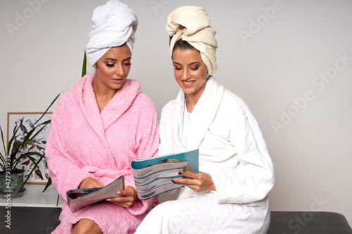 Two happy young women in bathrobes and wrapped towel on their heads relax and read beauty magazines in a beauty studio