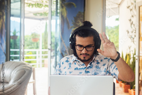 Young Latino male college student with headset, raising his hand in class in virtual classroom in front of laptop by zoom. video conference call at home.
