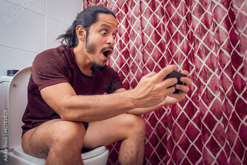 Young latin man smiling on the toilet using a smart phone at the bathroom. Man using the smartphone to play video games at the bathroom sitting on the toilet.