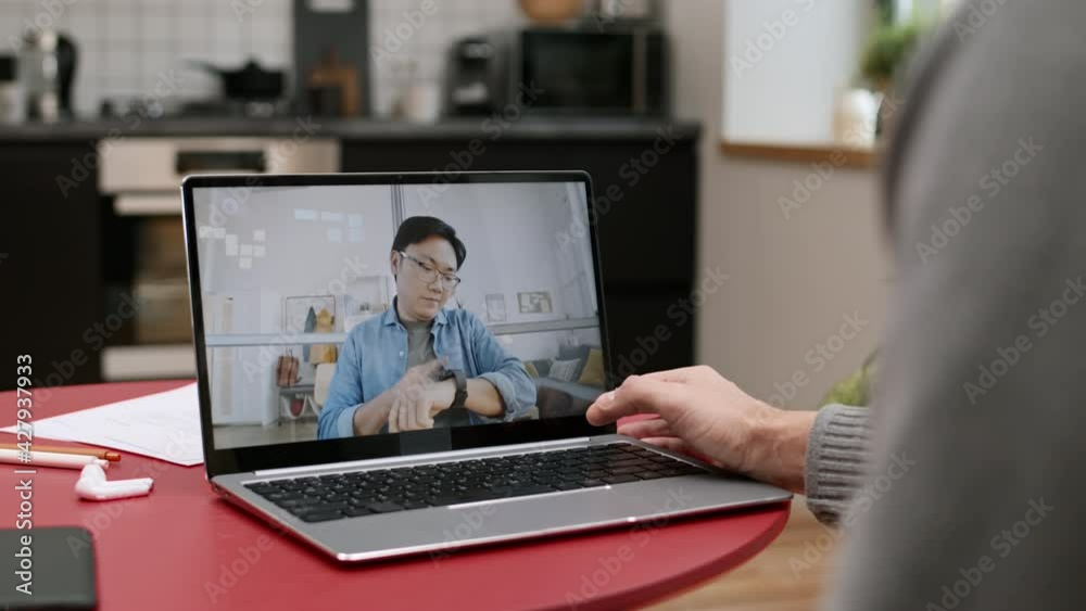 Over shoulder of unrecognizable person sitting at table with portable computer with running video conference on of young Asian man talking Over shoulder of unrecognizable person sitting at table with portable computer with running video conference on of young Asian man talking
