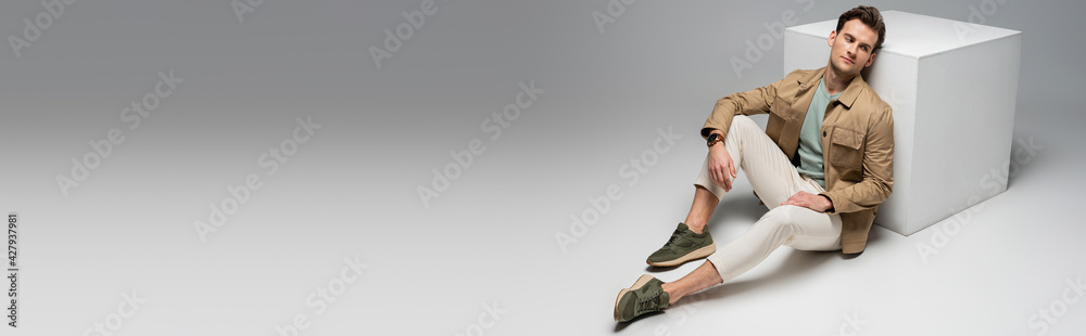 full length of stylish man posing while leaning on white cube on grey, banner full length of stylish man posing while leaning on white cube on grey, banner
