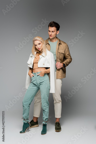 full length of model in jacket posing with hands in pockets near man on grey