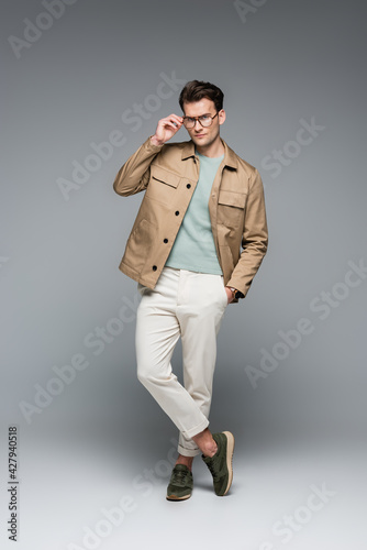 full length of trendy man in stylish outfit posing with hand in pocket on grey