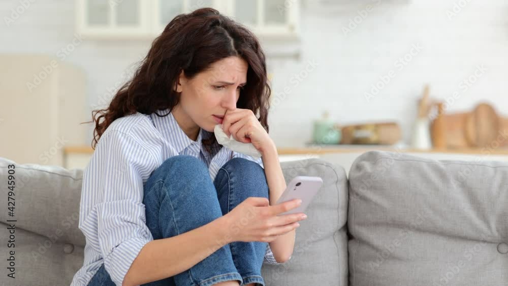 Young woman cry reading bad news email, break up message from ex-boyfriend on smartphone sitting lonely on couch in living room. Depressed unhappy female desperate of heartbreak sms on mobile phone