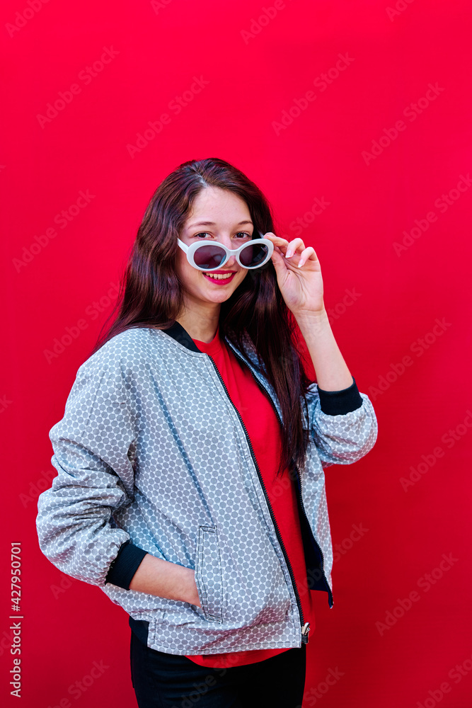 Obraz premium American flat portrait of a smiling young brunette girl taking off her sunglasses on flat red background