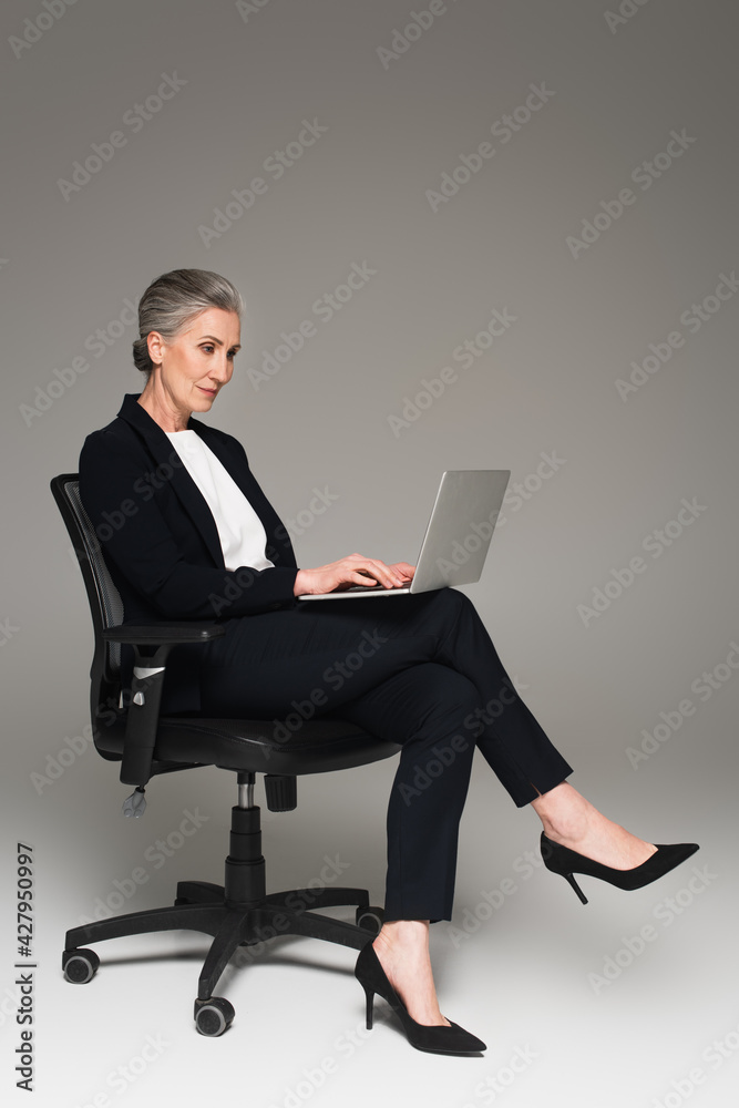 Mature businesswoman using laptop on office chair on grey background Mature businesswoman using laptop on office chair on grey background