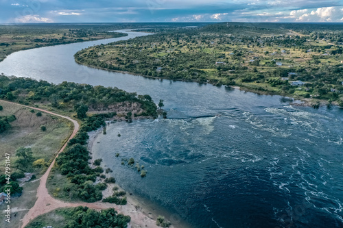 Wide panoramic view of Zambezi River in Zambia showing rapids, gravel road on riverbank and lush vegetation.